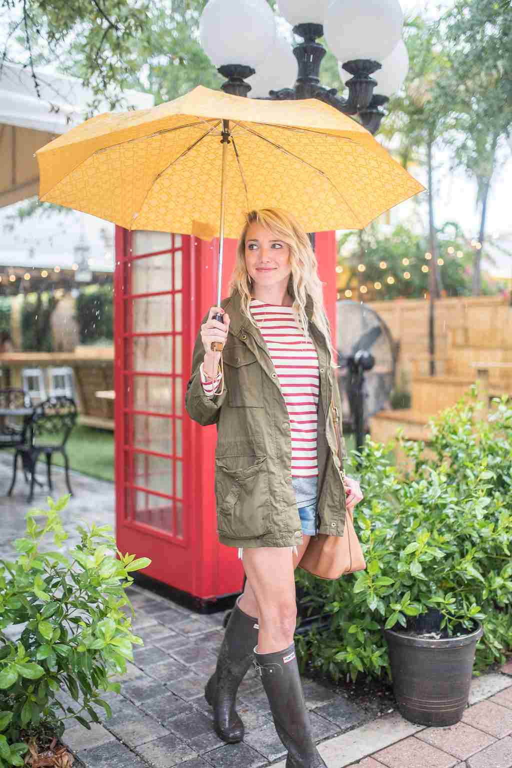 Shorts, botas de borracha, camiseta e jaqueta cáqui como traje e guarda-chuva amarelo como proteção da chuva