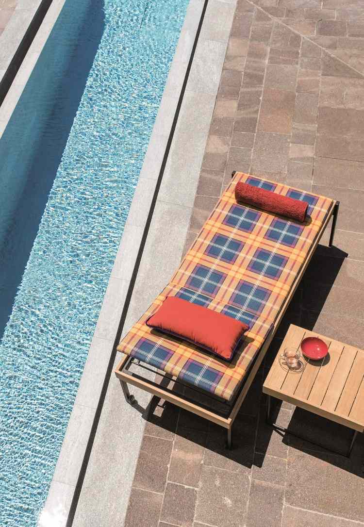 Espreguiçadeira com almofadas coloridas ao lado da piscina com mesinha de madeira ao ar livre