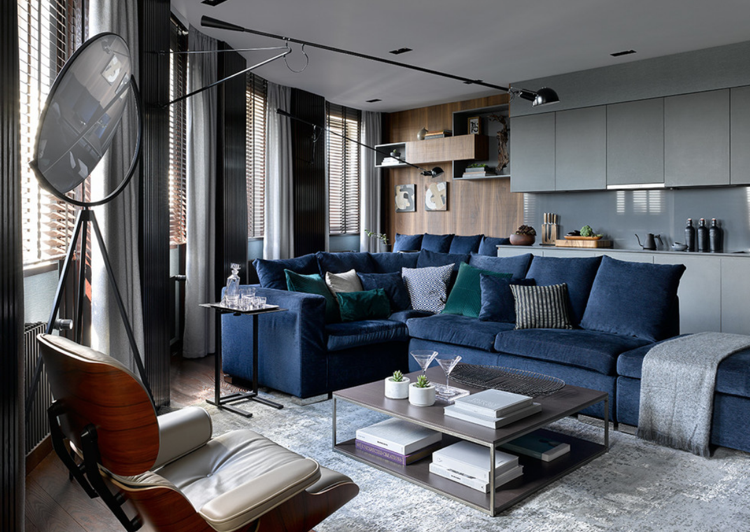 Sala de estar azul escuro com sofá de canto cinza com cortinas e cozinha compacta